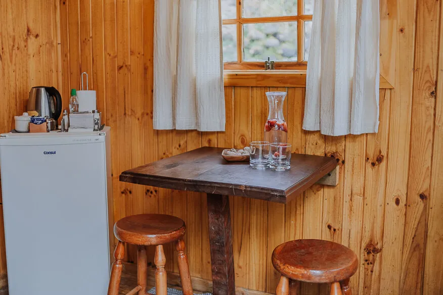 Rincón de comedor de la cabaña Eucaliptus con mesa de madera, piso alfombrado y ventana al bosque